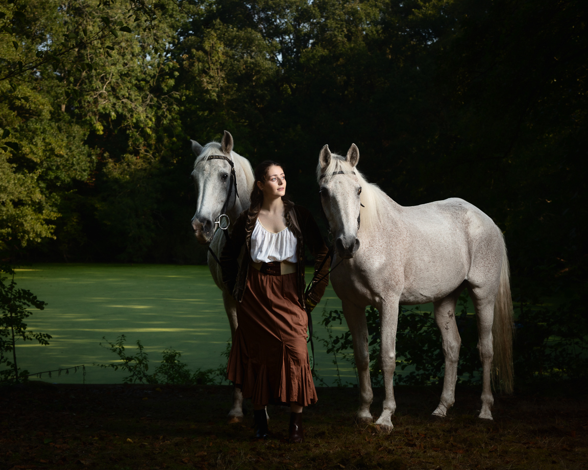 Woman in period dress standing with two white horses by water
