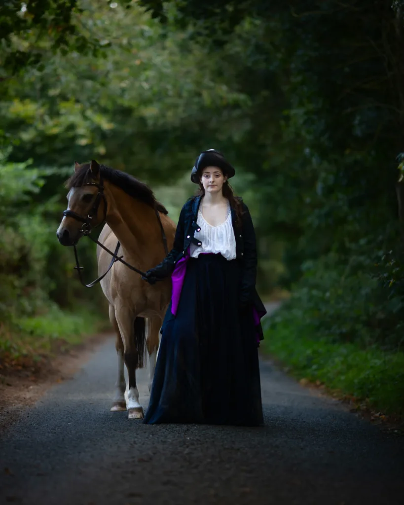 Woman in vintage dress walking with brown horse countryside lane