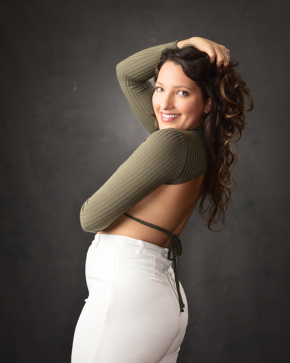 Woman in khaki top and white trousers posing against grey background
