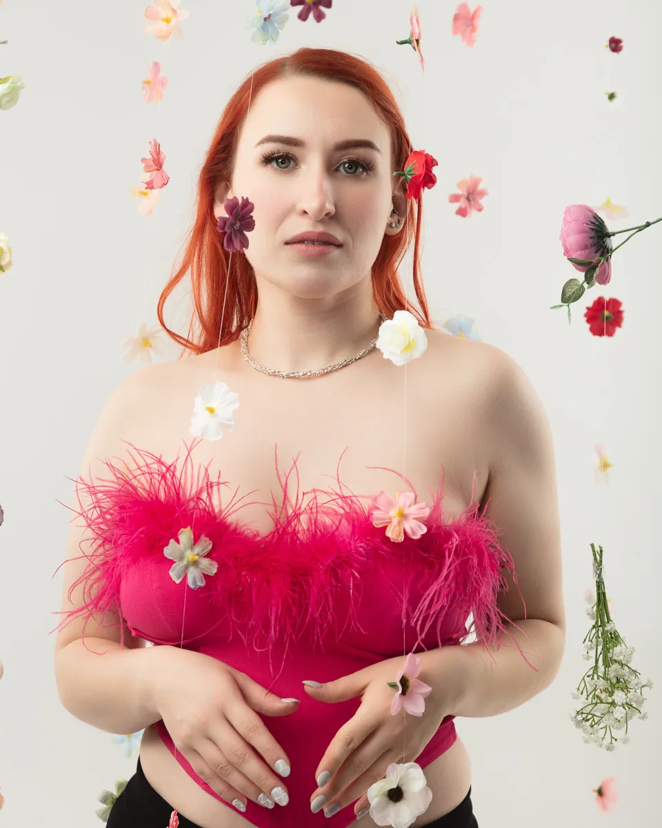 Woman with red hair wearing pink feathered top surrounded by flowers