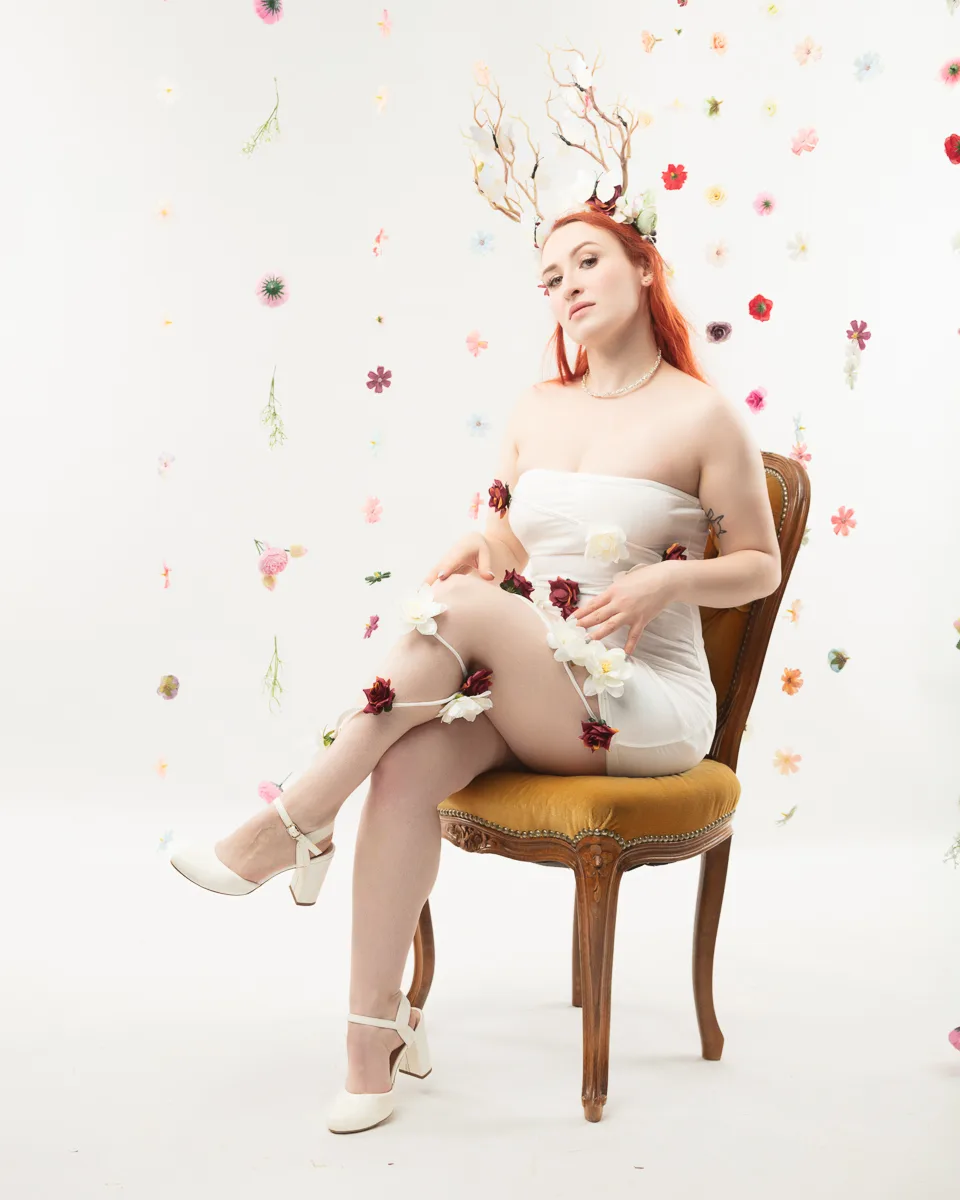 Woman with red hair wearing white dress sitting on wooden chair surrounded by floating flowers