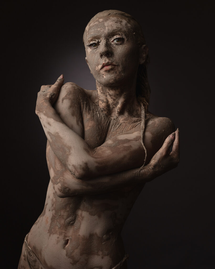 Person covered in mud, embracing themselves, dark background.