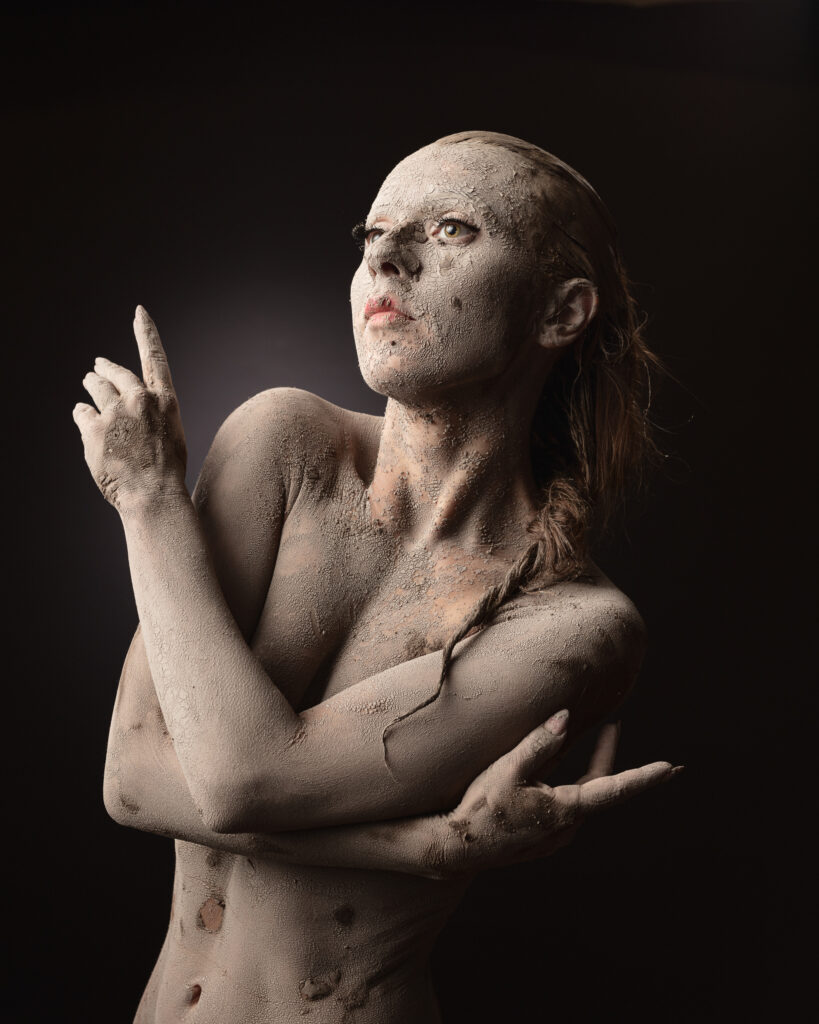 Fine art studio portrait, model covered in pale clay-like body paint against a dark background