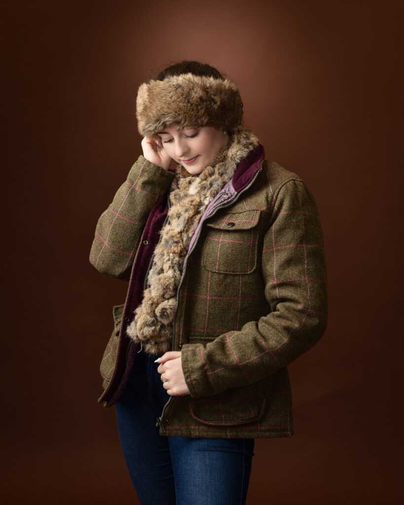 Country-style studio portrait of Sophia in a tweed jacket with fur-trim scarf, looking down while adjusting her fur hat