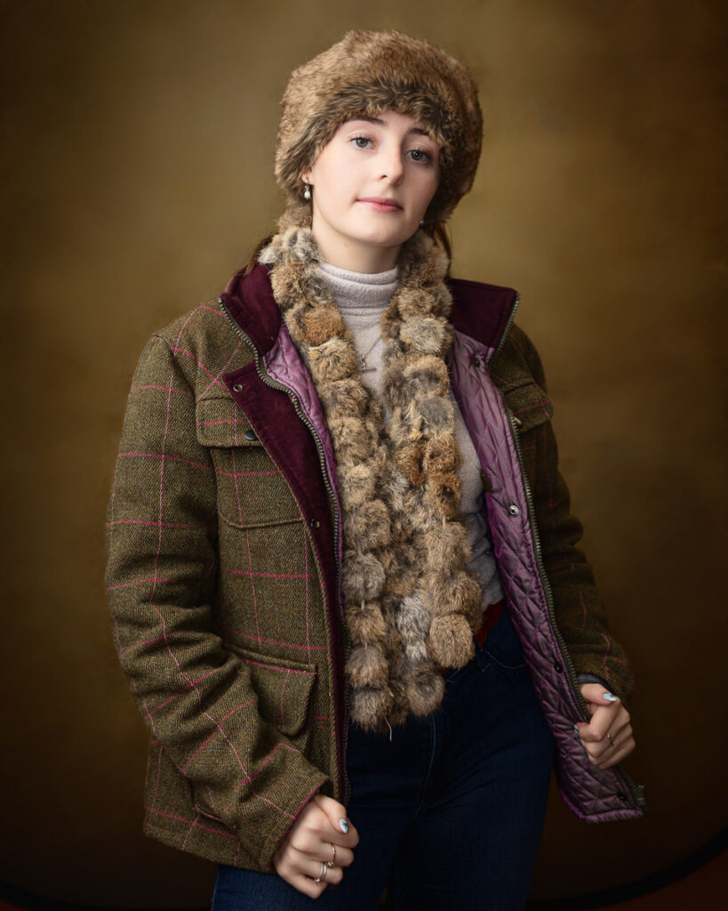 Country-style studio portrait of Sophia in a tweed jacket, fur hat and scarf, hands at the jacket zip
