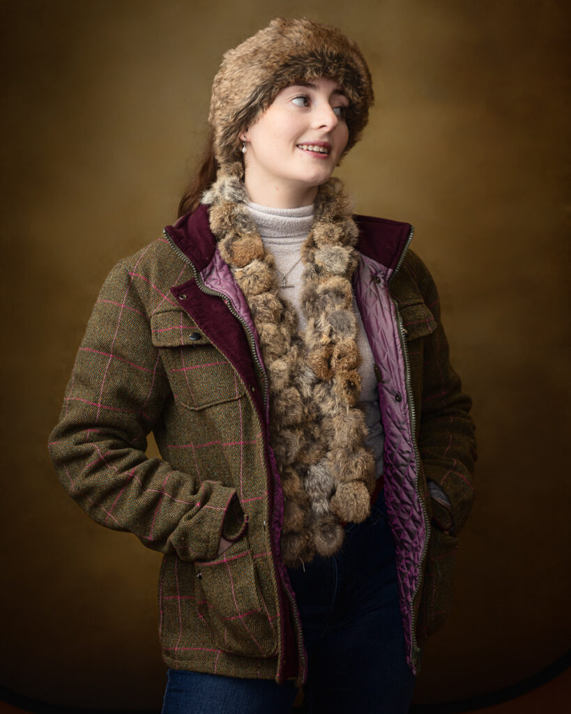 Country-style studio portrait of Sophia in a tweed jacket, fur hat and scarf, smiling with hands in pockets