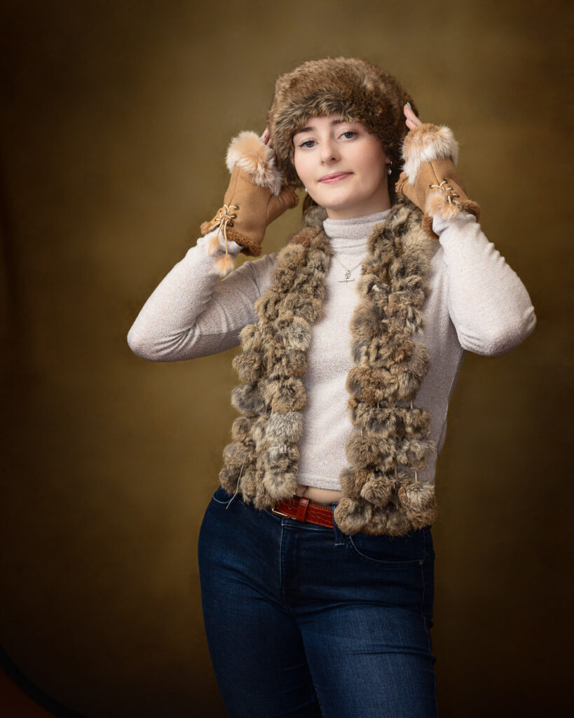 Country-style studio portrait of Sophia in a fur hat and mitts with a pom-pom trimmed gilet over a cream jumper and jeans