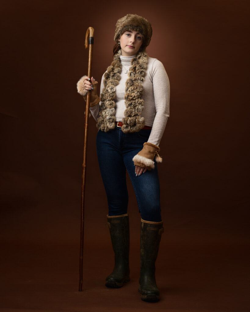 Studio portrait of Sophia dressed as a shepherdess, in a fur hat and scarf holding a wooden crook, side angle
