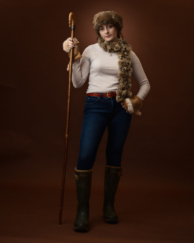 Studio portrait of Sophia dressed as a shepherdess, in a fur hat and scarf holding a wooden crook, with green wellington boots