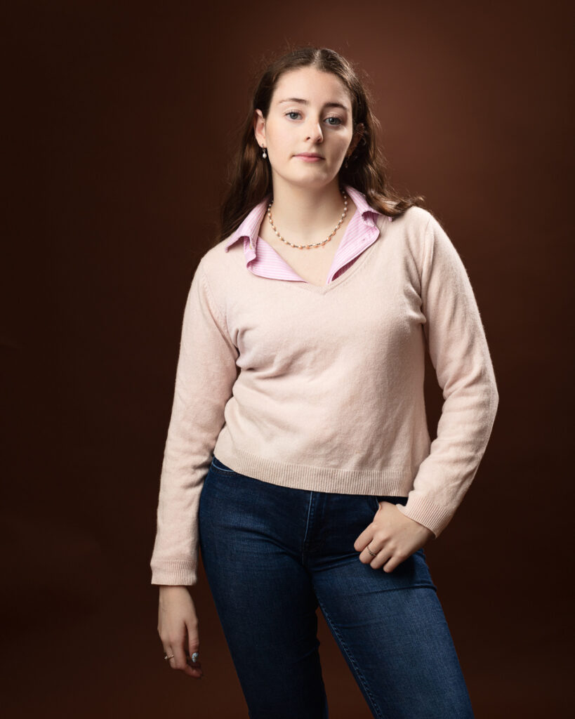 Studio portrait of Sophia in a pink jumper over a pink striped shirt and jeans, direct gaze