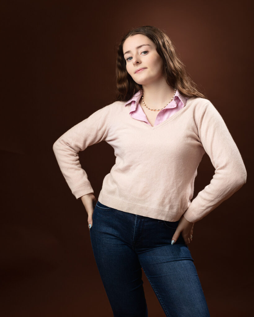 Studio portrait of Sophia in a pink jumper over a pink striped shirt and jeans, hands on hips