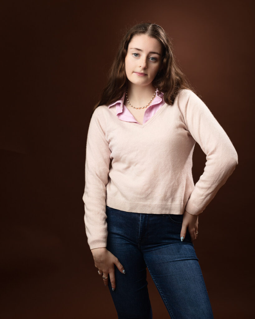 Studio portrait of Sophia in a pink jumper over a pink striped shirt and jeans, hand behind her back
