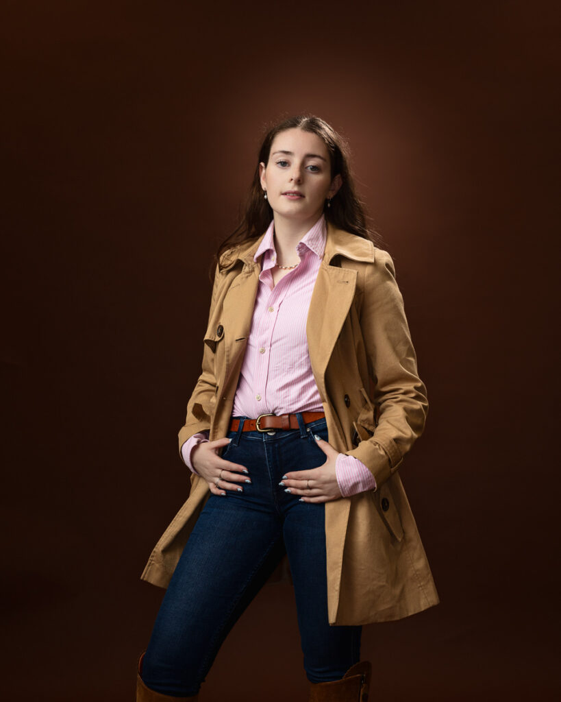 Studio portrait of Sophia in a tan trench coat over a pink striped shirt and jeans, front on