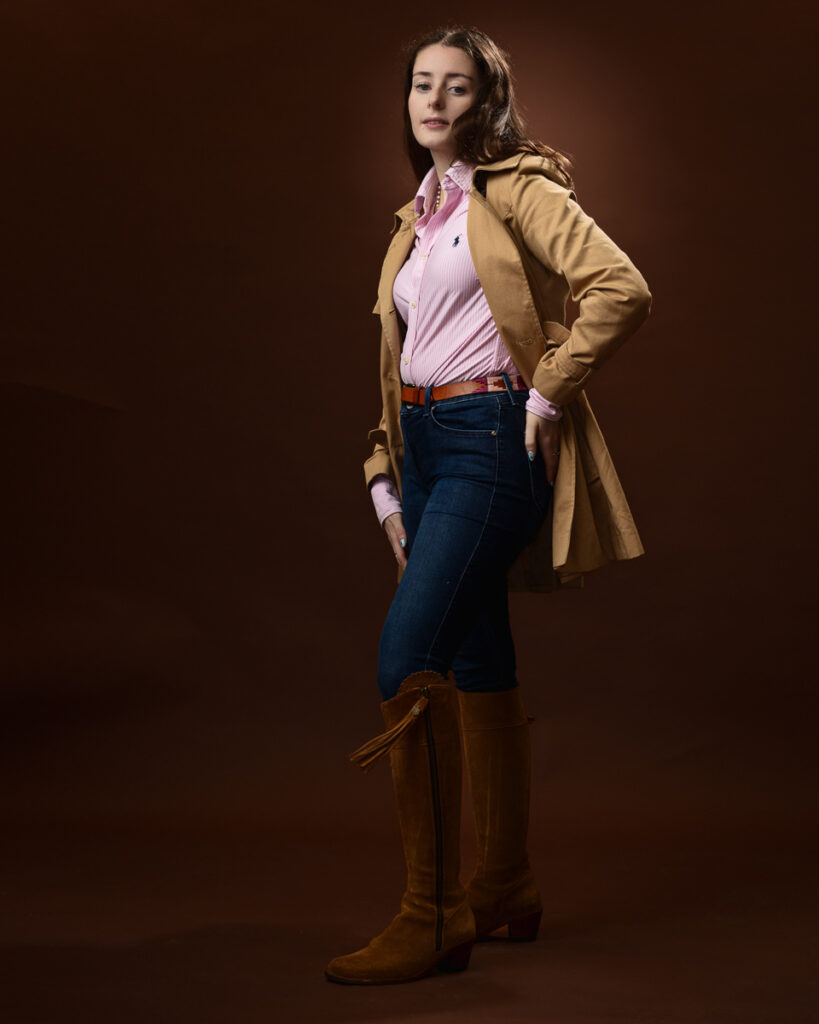 Studio portrait of Sophia in a tan trench coat over a pink striped shirt and jeans, hand on hip