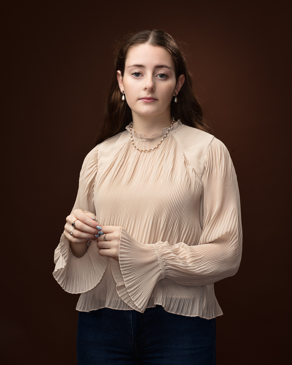 Studio portrait of Sophia in a cream pleated blouse with a pearl necklace, hands clasped together