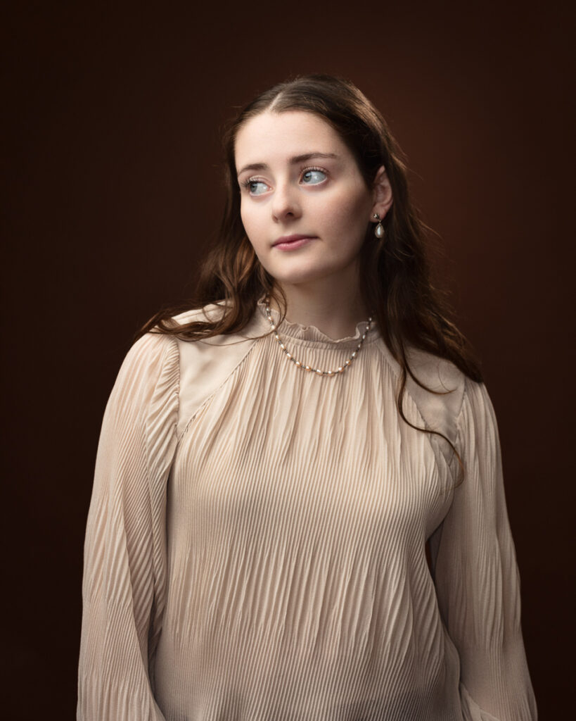 Studio portrait of Sophia in a cream pleated blouse with a pearl necklace, looking to the side