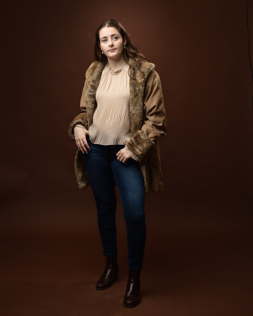 Studio portrait of Sophia in a tan faux-fur trimmed coat over a cream blouse and jeans, hands in pockets