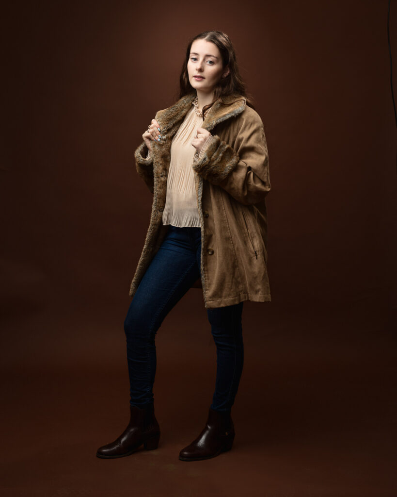 Studio portrait of Sophia in a tan faux-fur trimmed coat, holding the lapels, jeans and boots