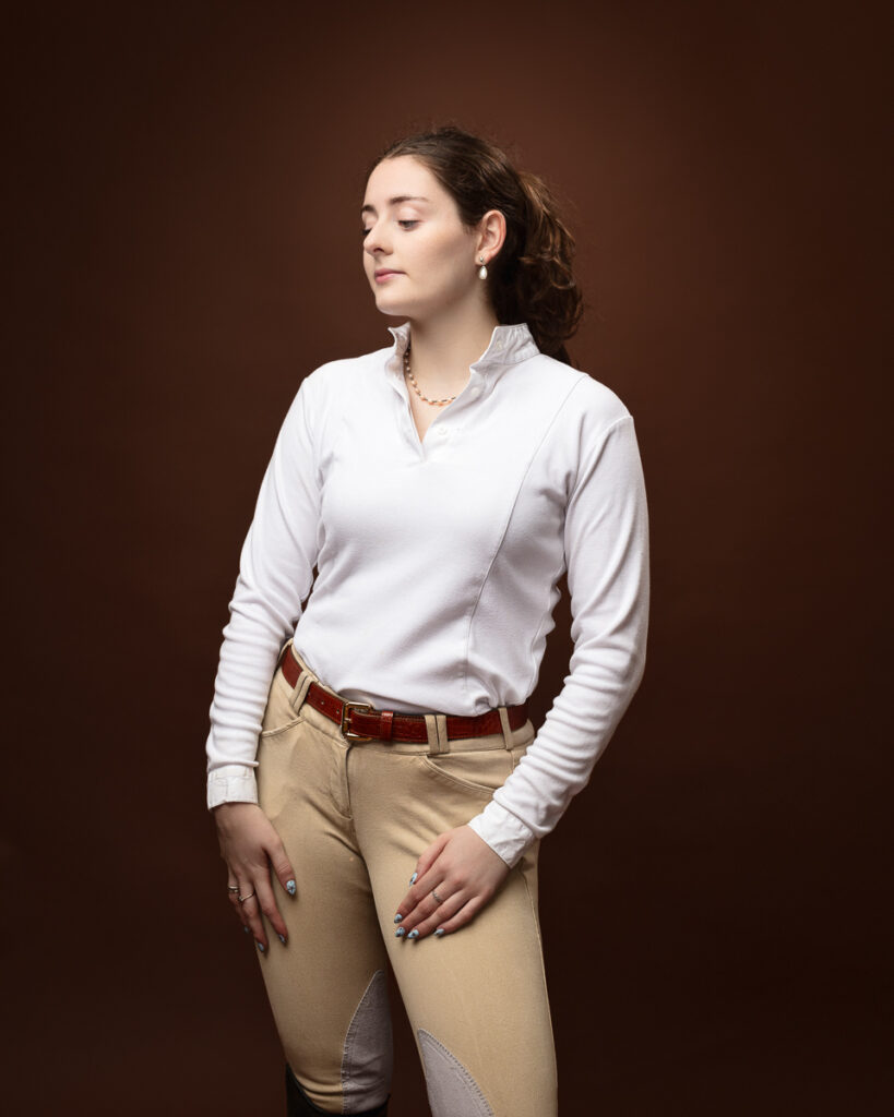 Equestrian-style studio portrait of Sophia in a white riding top and tan jodhpurs, looking down