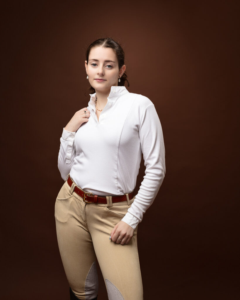 Equestrian-style studio portrait of Sophia in a white riding top and tan jodhpurs, hand at the collar