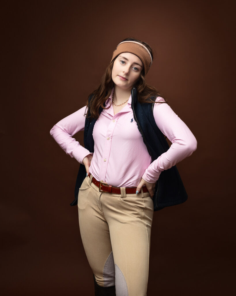 Equestrian-style studio portrait of Sophia in a navy gilet over a pink shirt with a headband, hands on hips