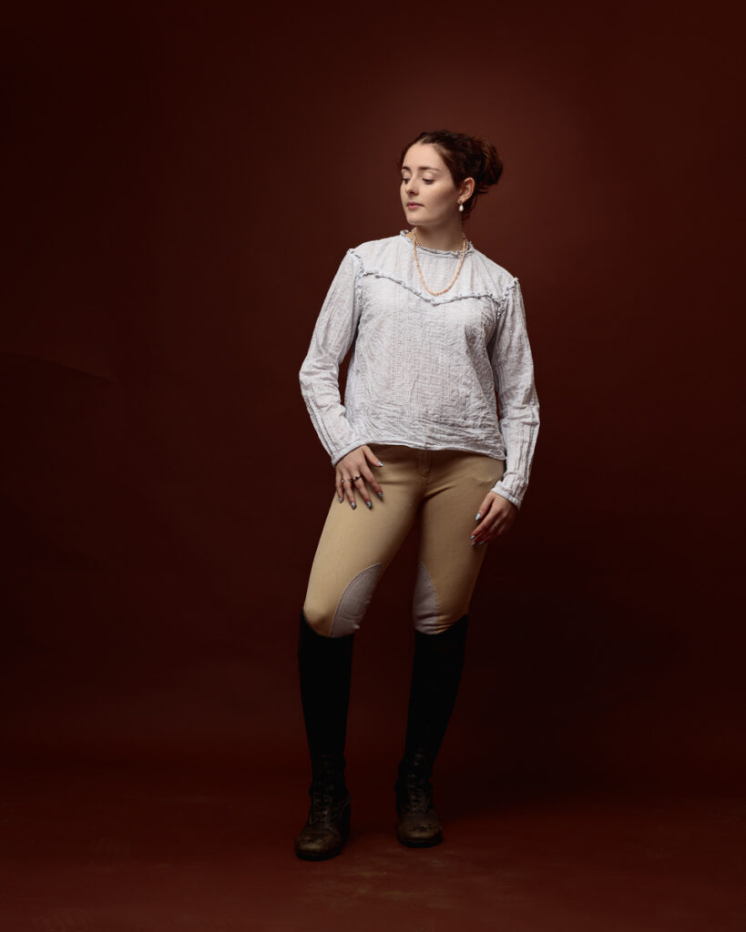Equestrian-style studio portrait of Sophia in a cream blouse and tan jodhpurs, looking down