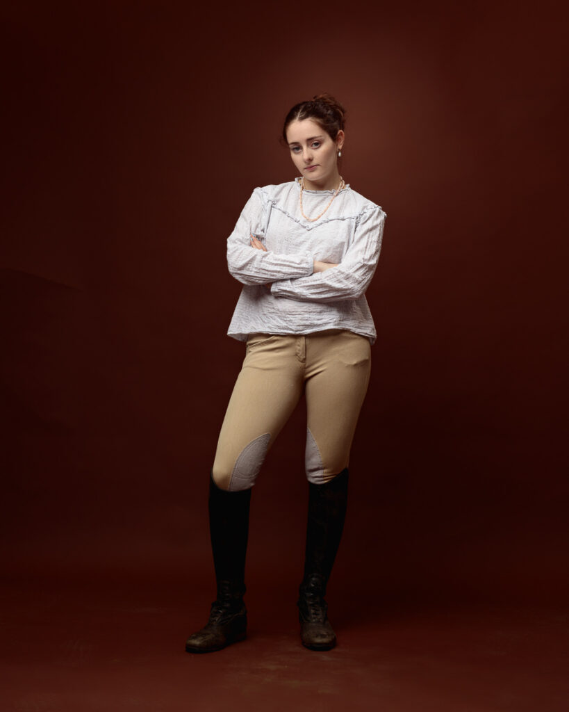 Equestrian-style studio portrait of Sophia in a cream blouse, tan jodhpurs and riding boots, arms crossed