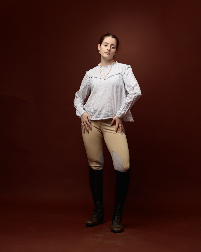 Equestrian-style studio portrait of Sophia in a cream blouse, tan jodhpurs and black riding boots, hands on hips