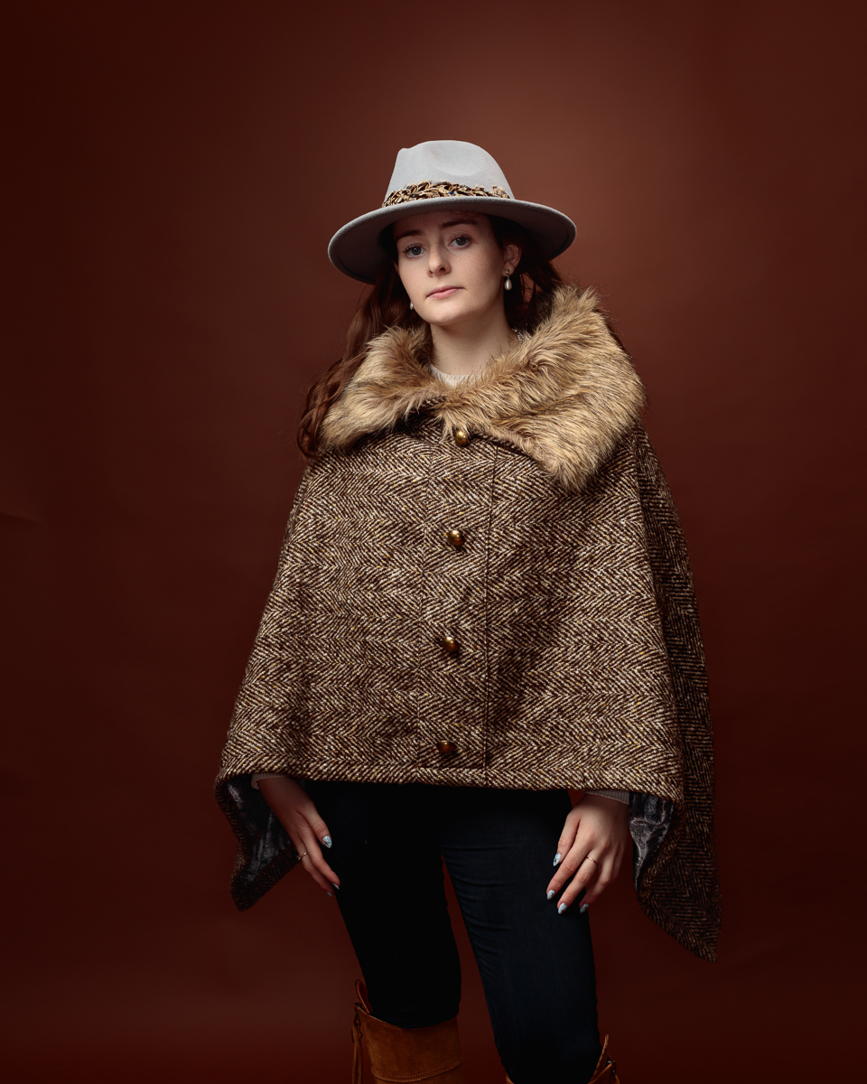 Studio portrait of Sophia in a tweed fur-trimmed cape and grey fedora, standing front on with knee-high boots