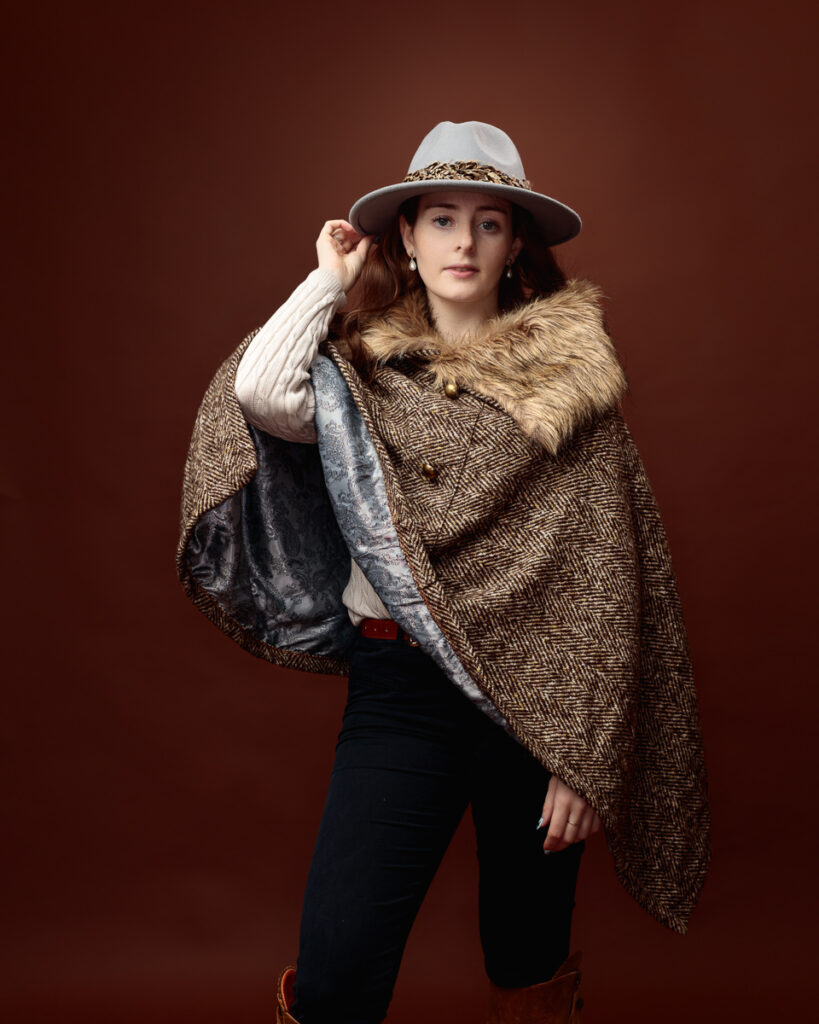 Studio portrait of Sophia in a tweed fur-trimmed cape and grey fedora, touching the hat brim with the cape open showing its lining