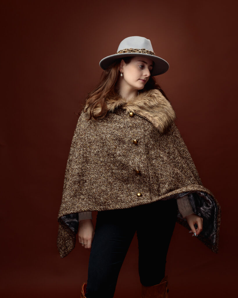 Studio portrait of Sophia in a tweed fur-trimmed cape and grey fedora, looking to the side with arm folded