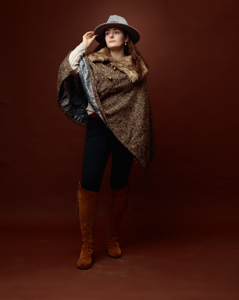 Studio portrait of Sophia in a tweed fur-trimmed cape and grey fedora, looking to the side with the cape flowing open