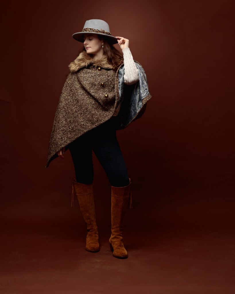 Studio portrait of Sophia in a tweed fur-trimmed cape and grey fedora, touching the hat brim and looking down