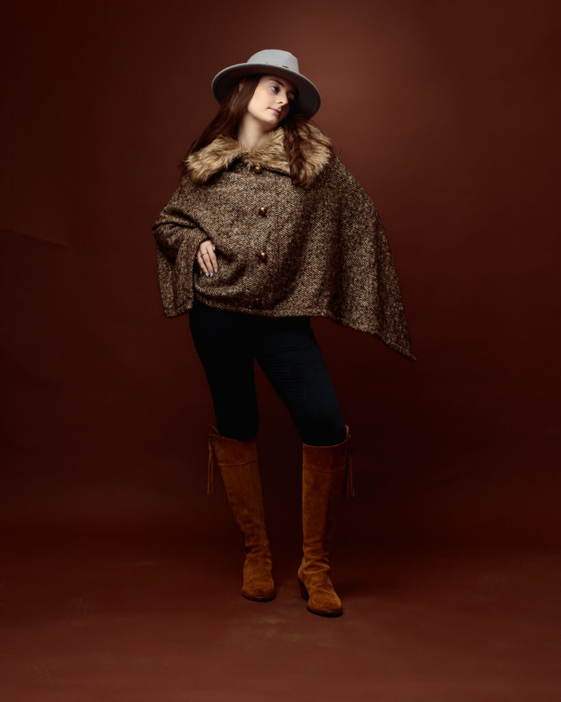 Studio portrait of Sophia in a tweed fur-trimmed cape and grey fedora, side profile with hand on hip