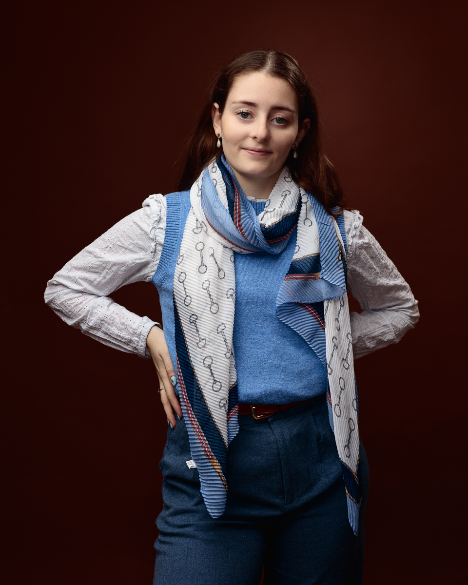 Studio portrait of Sophia in a blue jumper and white blouse with a horse-bit print scarf, hand on hip