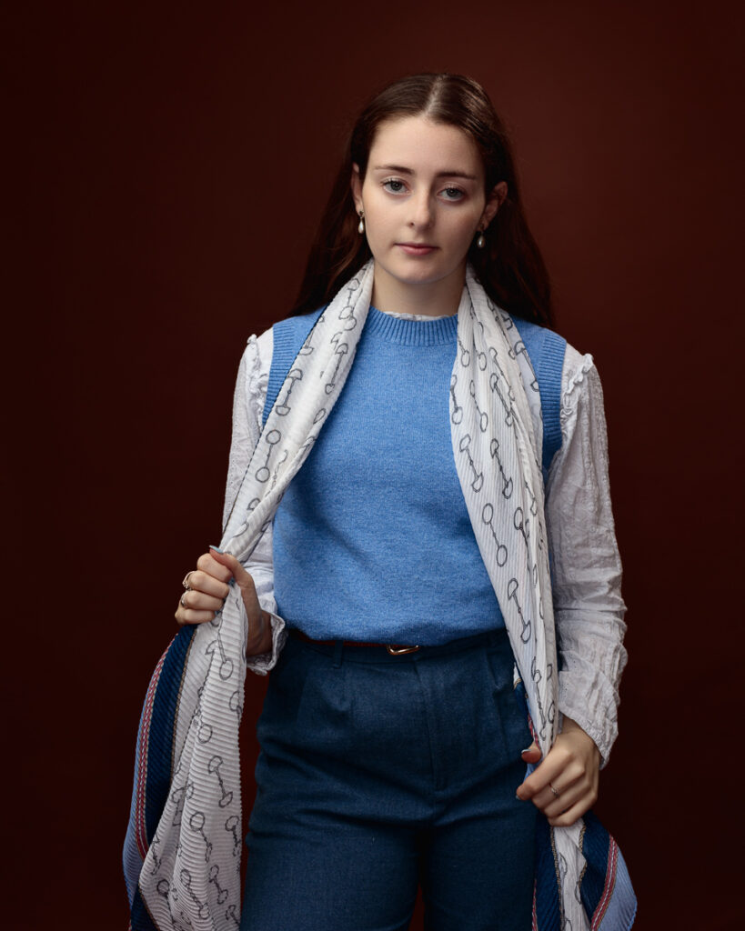 Studio portrait of Sophia in a blue jumper with a horse-bit print scarf, holding the scarf ends