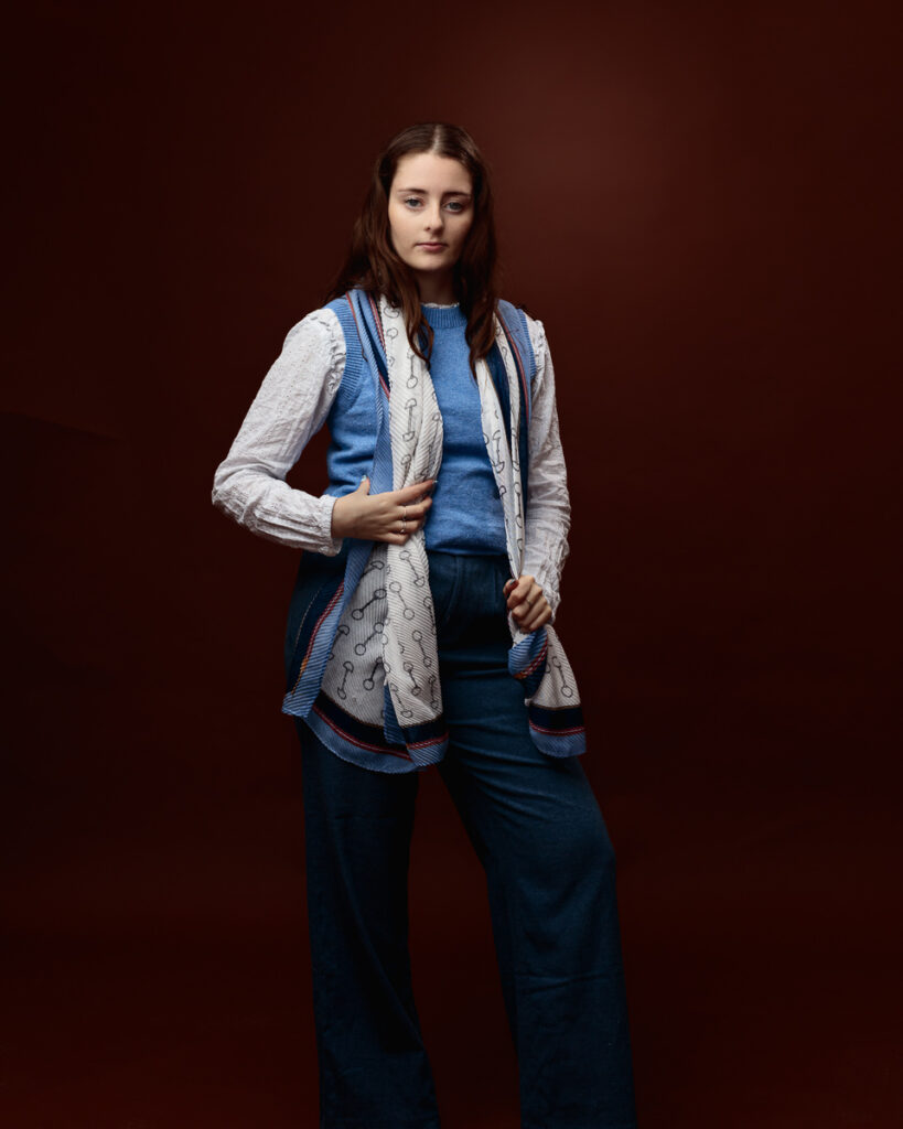 Studio portrait of Sophia in a blue jumper and white blouse with a horse-bit print scarf, hand near the scarf