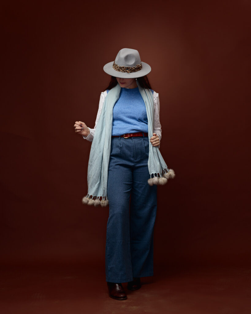 Studio portrait of Sophia in a grey fedora tilted down, wearing a mint pom-pom trimmed scarf and wide-leg jeans