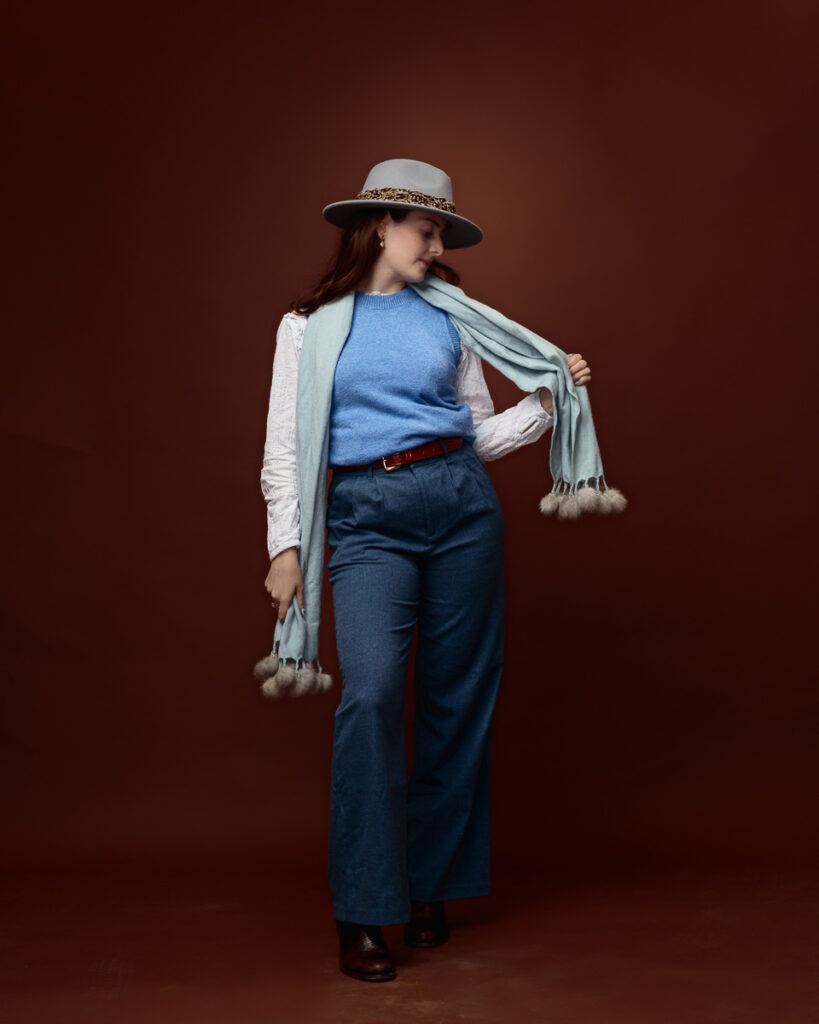 Studio portrait of Sophia in a grey fedora with a mint pom-pom scarf over her shoulder, walking in wide-leg jeans