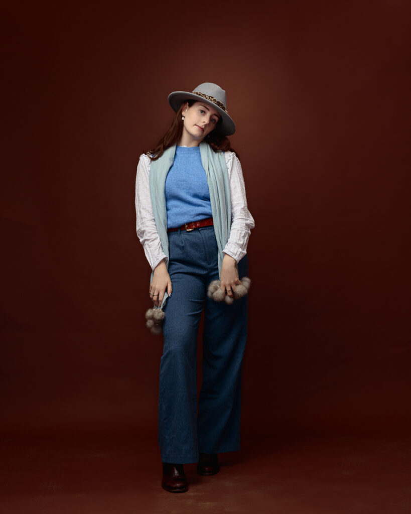 Studio portrait of Sophia in a grey fedora, blue jumper and wide-leg denim trousers, with a pom-pom trimmed scarf