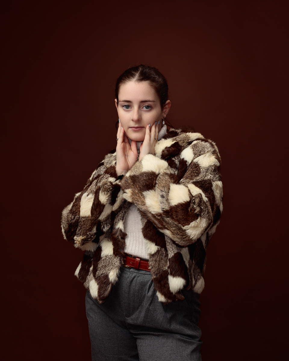 Studio portrait of Sophia in a patchwork faux fur coat, hands framing her face