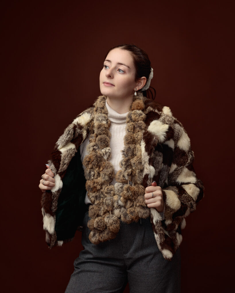 Studio portrait of Sophia in a patchwork faux fur coat, looking to the side while holding the lapels open
