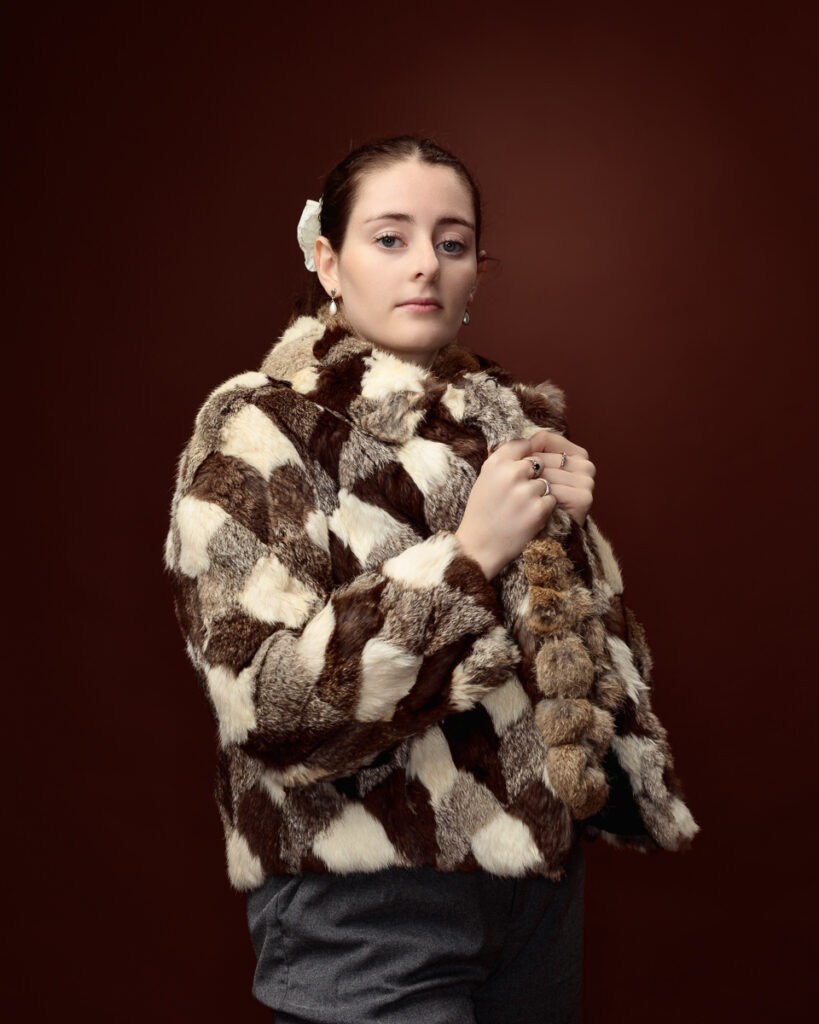 Studio portrait of Sophia in a patchwork faux fur coat with pom-pom trim, side angle holding the collar