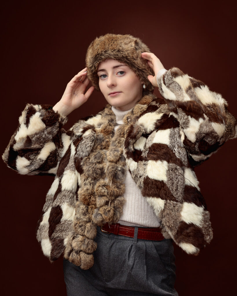 Studio portrait of Sophia in a patchwork faux fur coat, adjusting her matching fur hat with both hands