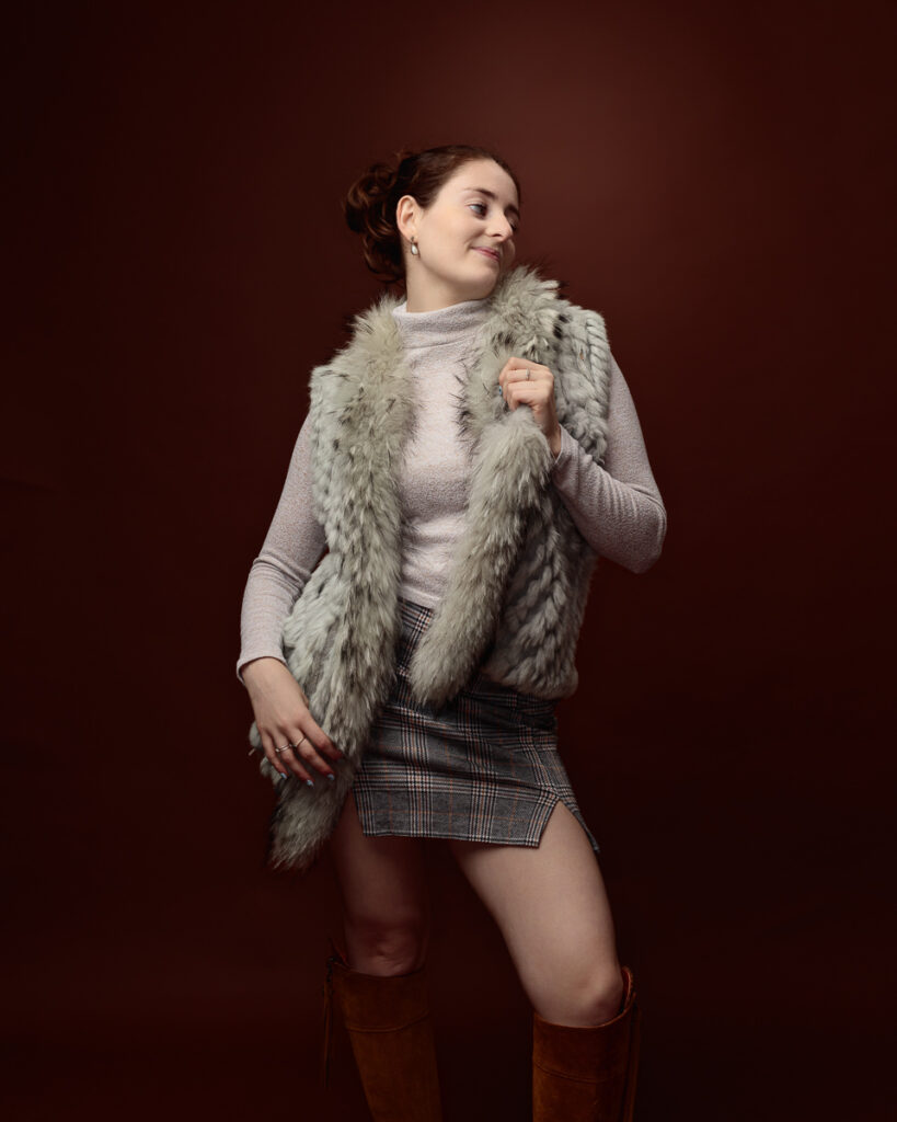 Studio portrait of Sophia in a sage green faux fur gilet and tartan mini skirt, looking to the side and smiling