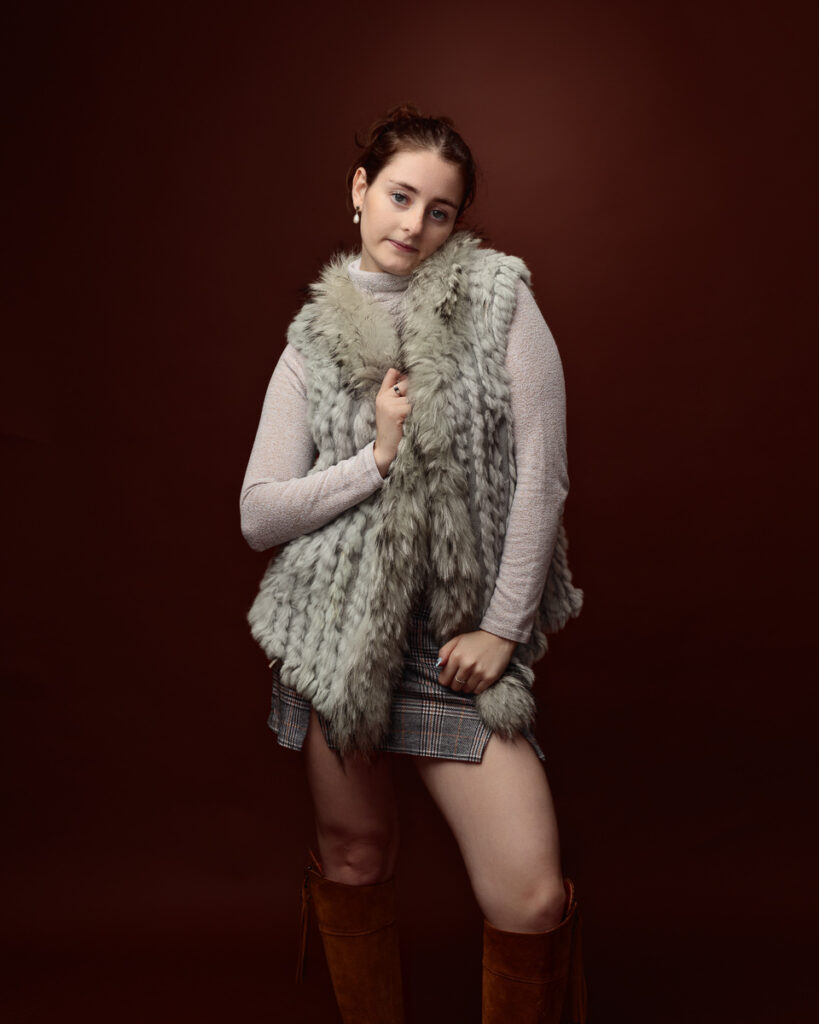 Studio portrait of Sophia in a sage green faux fur gilet, cream jumper and tartan mini skirt, hand at the collar