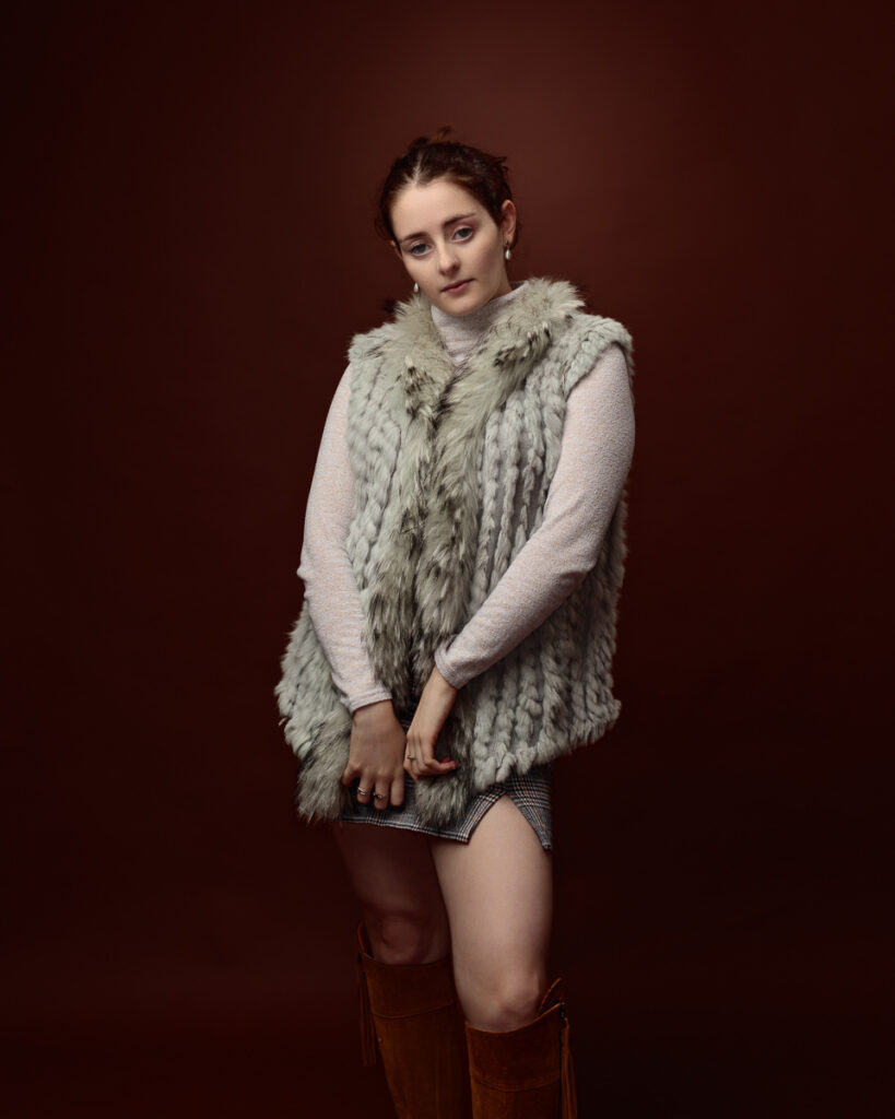 Studio portrait of Sophia in a sage green faux fur gilet over a cream jumper and tartan mini skirt, hands together
