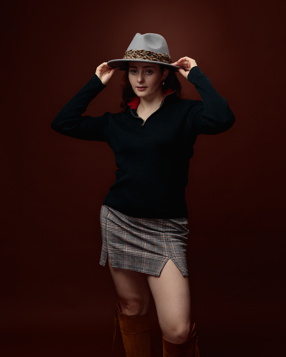 Studio portrait of Sophia in a black zip-neck jumper and tartan mini skirt, holding the brim of a grey fedora