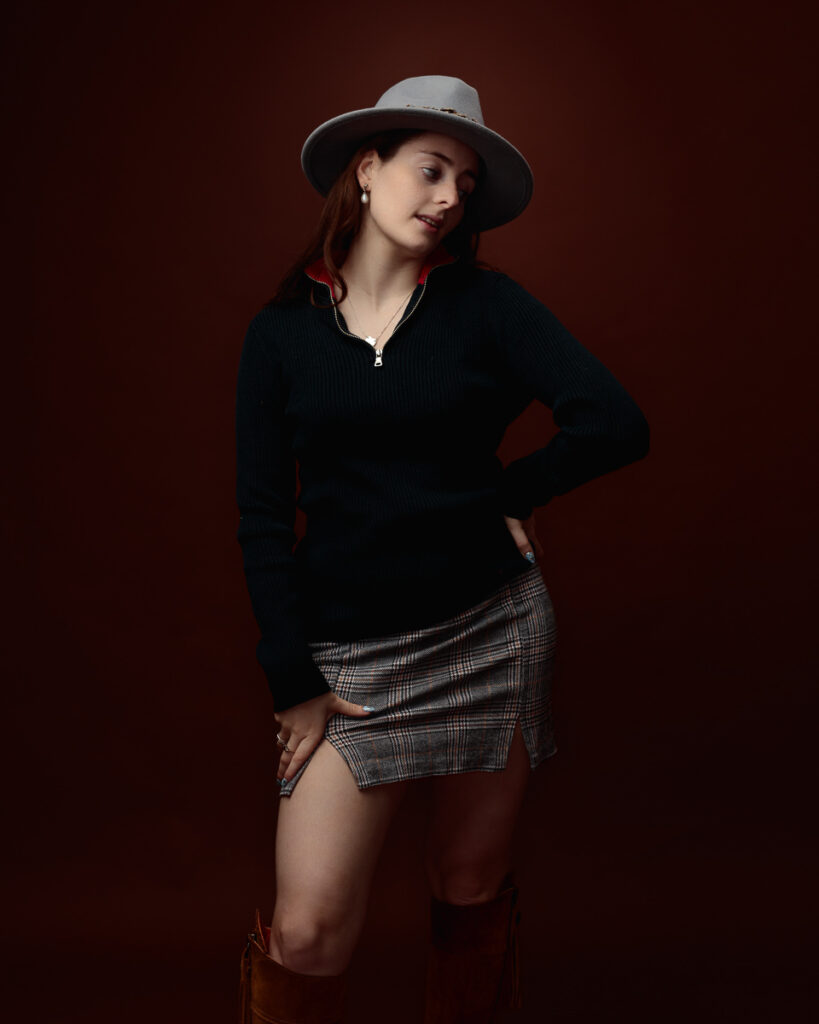 Studio portrait of Sophia in a black zip-neck jumper, tartan mini skirt and grey fedora, looking to the side