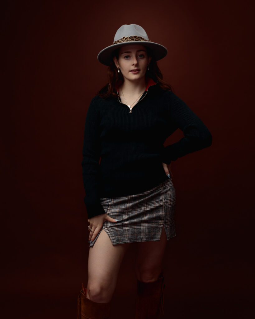 Studio portrait of Sophia in a black zip-neck jumper, tartan mini skirt and grey fedora, hand on hip